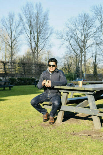 Abhishek S Chauhan sitting on a picnic bench in a park, holding a drink and wearing sunglasses on a sunny day.