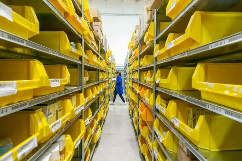 Worker in blue uniform walking through warehouse aisle with yellow plastic bins on metal shelves.