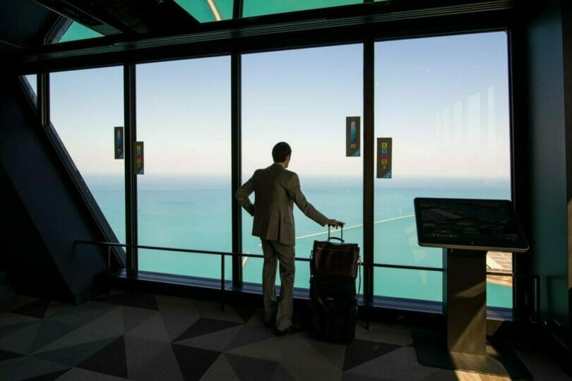 Man in a suit with a suitcase looks out from a tall building window at a vast body of water on a clear day.