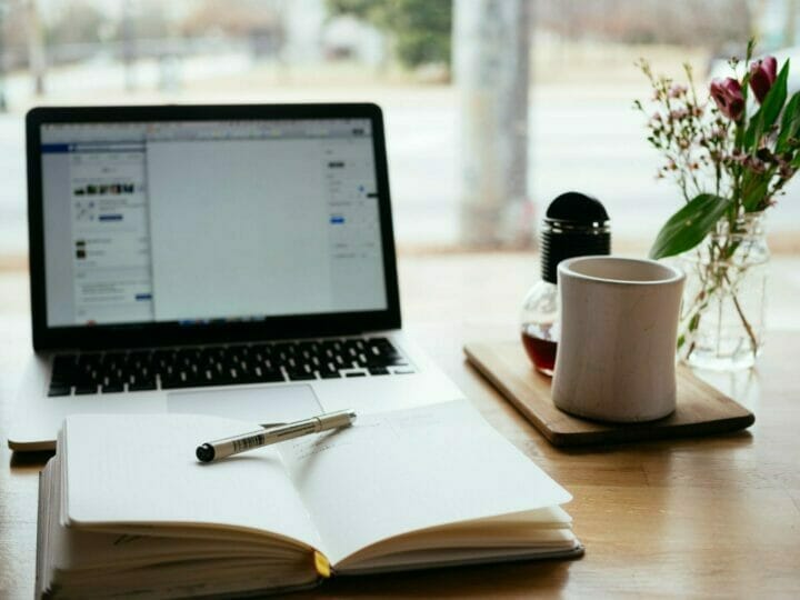 Open laptop on a wooden table with a notebook, pen, coffee cup, glass jar, and vase with flowers beside a window.