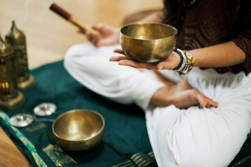 Person sitting cross-legged on the floor holding a singing bowl, with other bowls and decorative items placed on a cloth.