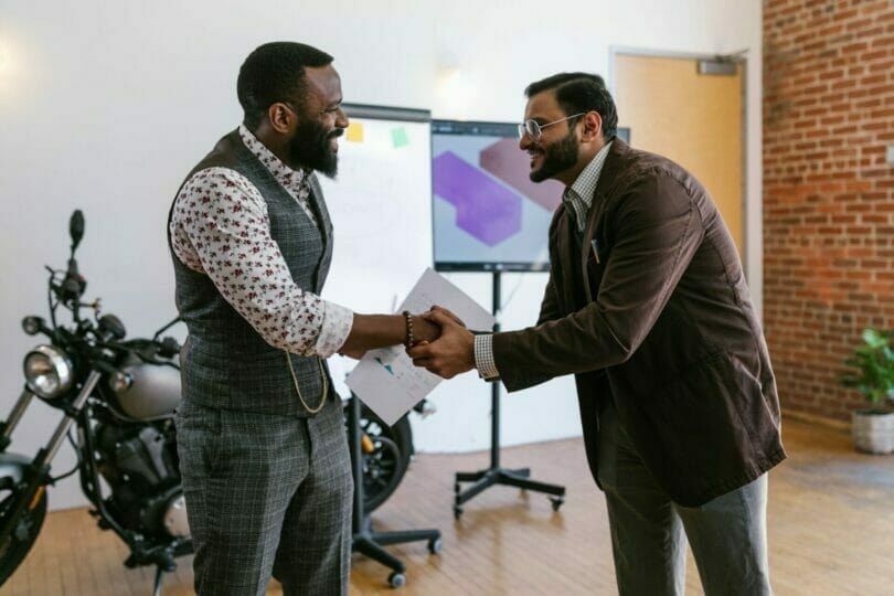 Two men in professional attire smiling and shaking hands in an office with a motorcycle and presentation board in the background.